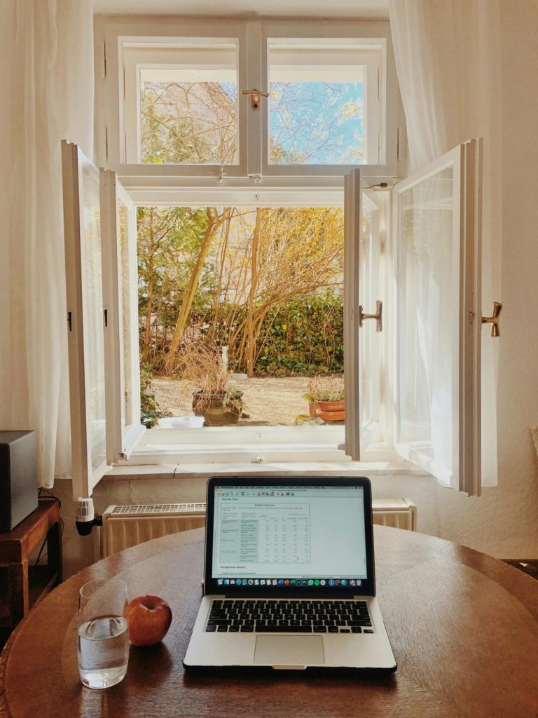 Ein Laptop auf einem Tisch mit einem Glas Wasser und einem Apfel, Blick auf einen Garten.