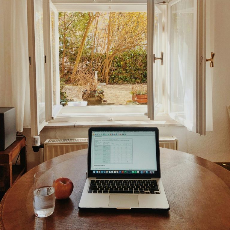 Schreibtisch mit Laptop, Glas Wasser und Apfel, Blick ins Freie durch offene Fenster.